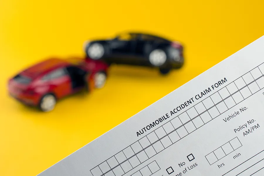 Close-up of an automobile accident claim form with two blurred toy cars in the background on a yellow surface, suggesting a collision and insurance paperwork assistance.